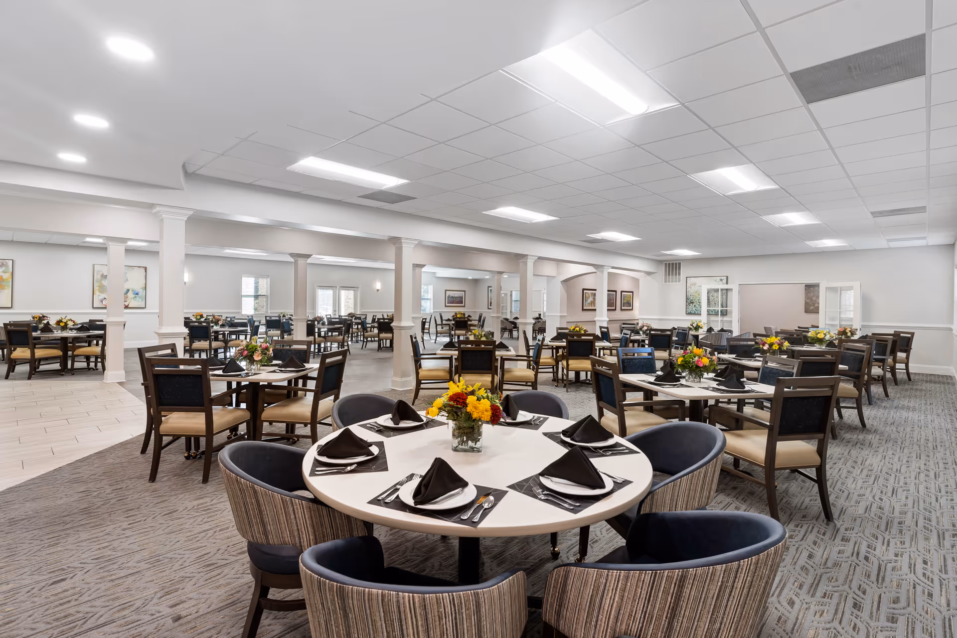 A spacious senior living dining room with multiple round and square tables set with black napkins, white plates, and silverware. Each table has a floral centerpiece. The room features a mix of carpeted and tiled flooring, white walls, and a ceiling with recessed lighting. Several framed artworks are visible on the walls.
