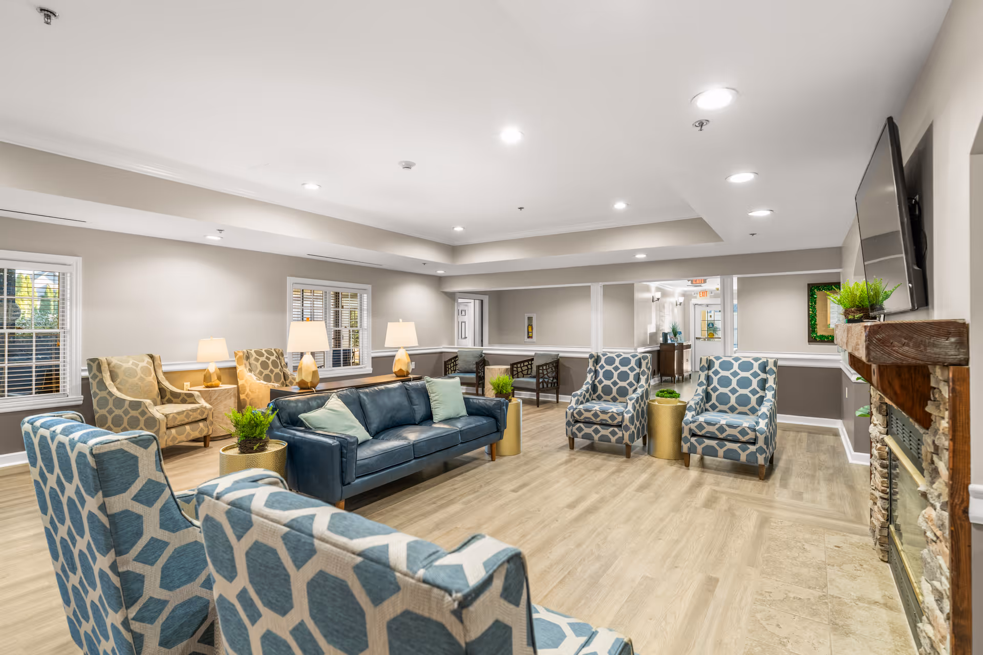 A spacious senior living facility common area with multiple patterned armchairs, a dark blue leather sofa with light green cushions, several side tables with lamps, potted plants, a stone fireplace with a wooden mantle, and a wall-mounted flat screen TV. The room has light wood flooring, beige walls with white trim, and recessed ceiling lights.