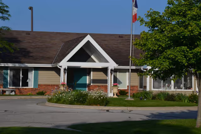 Front entrance of a single-story retirement residence with a gabled portico, flagpole, flower beds, and surrounding landscaping.
