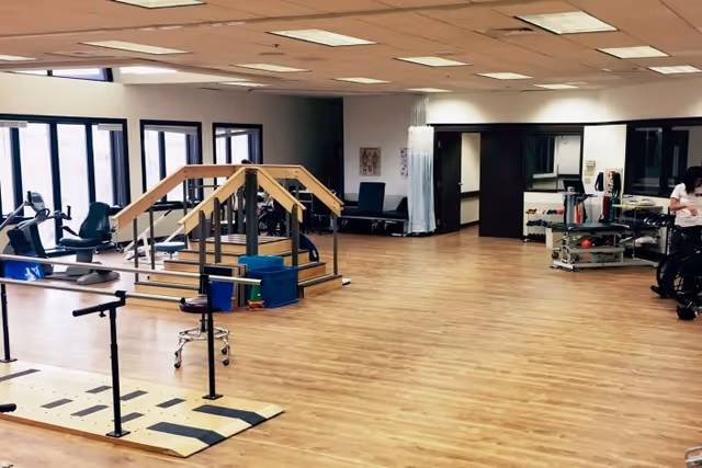 A spacious rehabilitation or physical therapy room with wooden flooring, parallel bars for walking exercises, a small set of stairs with handrails, exercise equipment, and a therapy table. Large windows line one side of the room allowing natural light in.