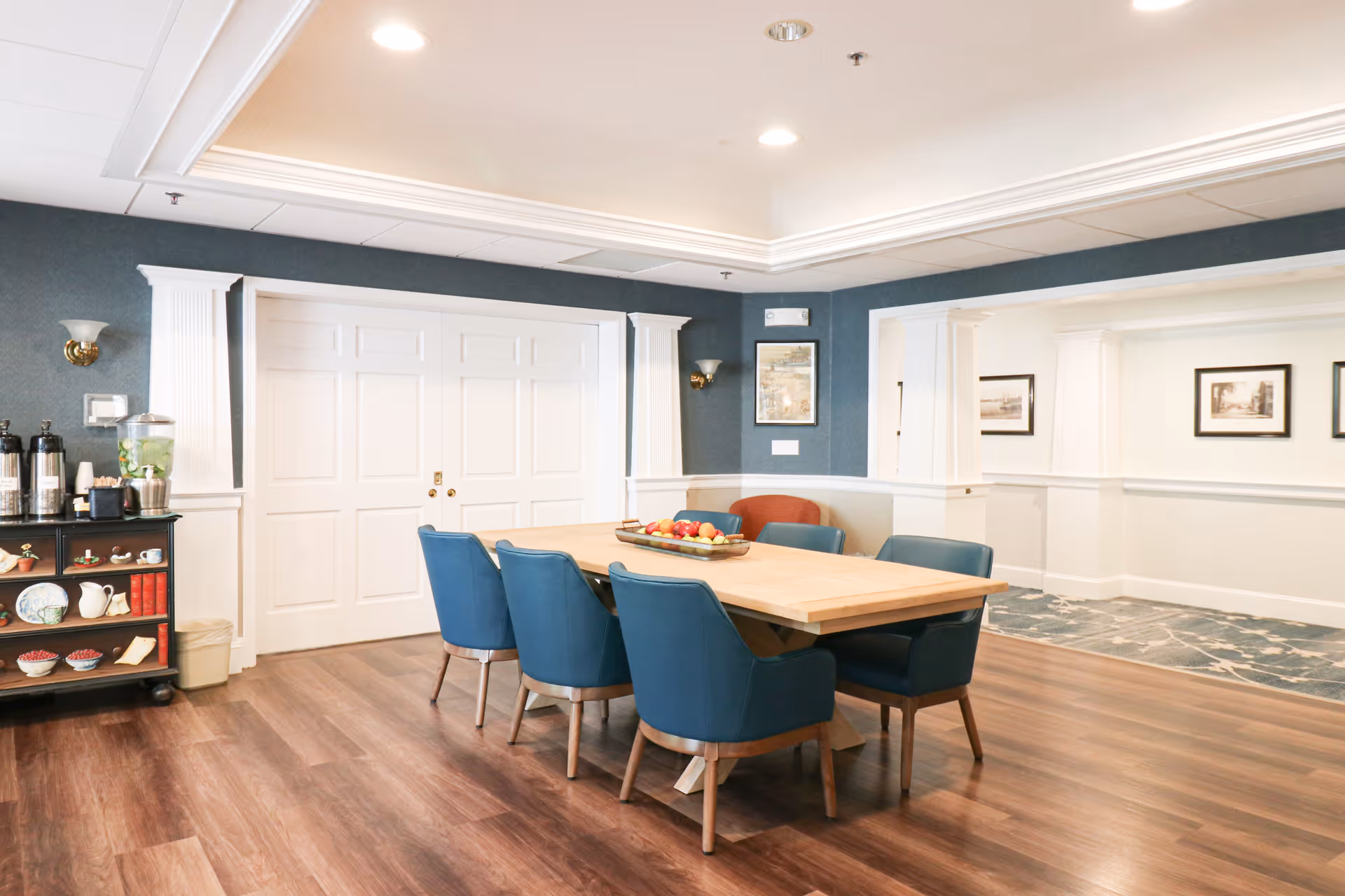 Bright communal dining area with a wooden table surrounded by blue chairs, a beverage cart, and white paneled walls.