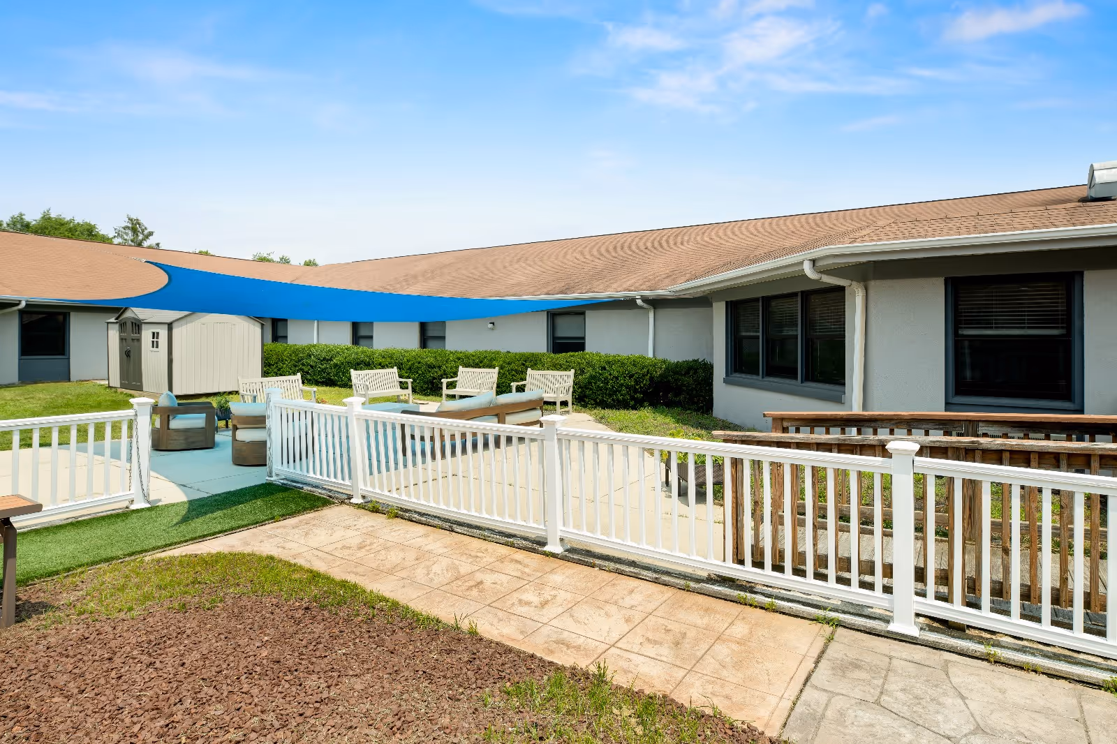 Outdoor patio area at Evergreen Post Acute with white railings, several chairs and tables under a large blue shade sail, surrounded by a single-story building with beige walls and brown roof, and some greenery.