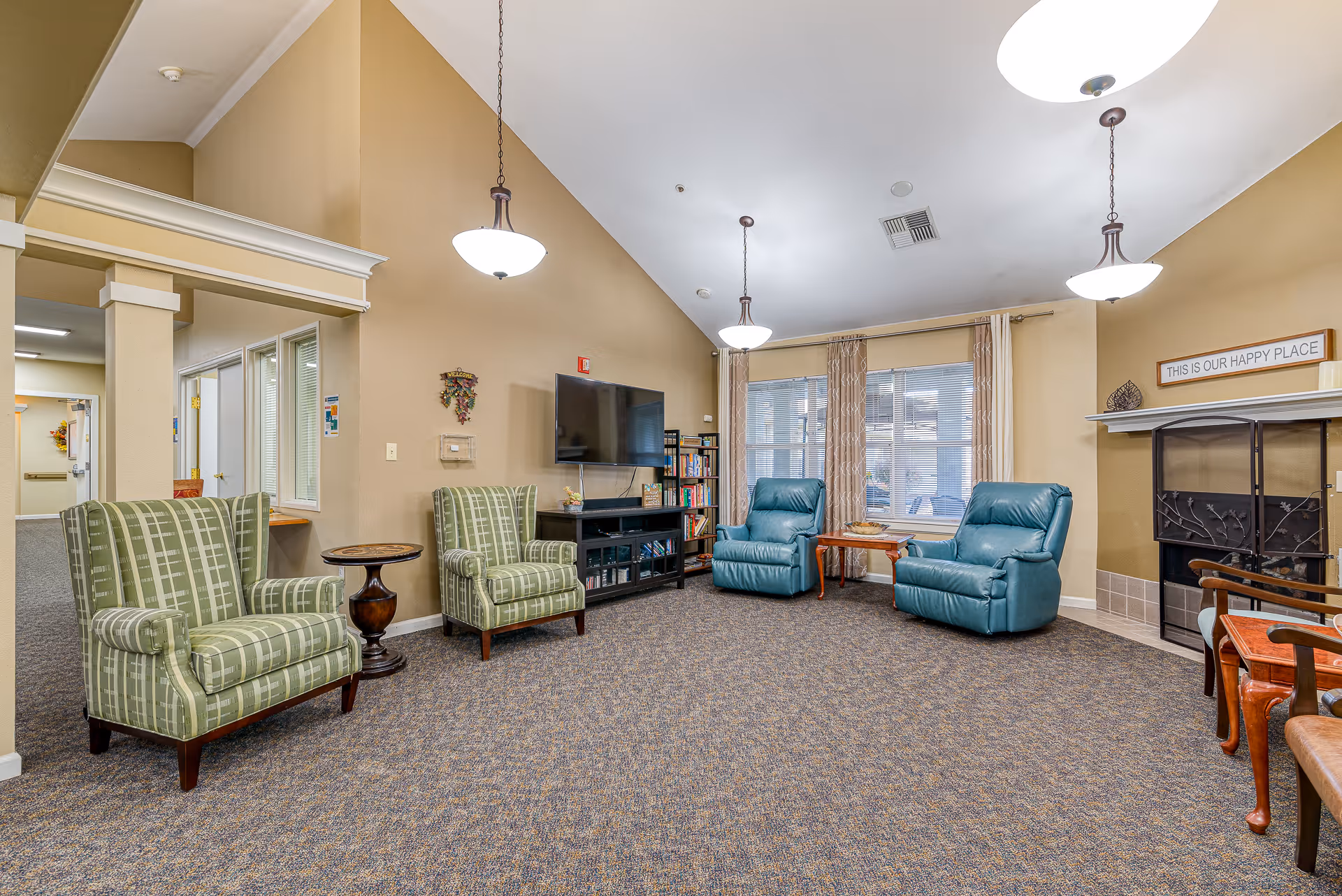 A cozy living room area in a senior living facility with beige walls and carpeted floor. The room features two green patterned armchairs, two blue recliners, a wooden side table, a TV mounted on the wall above a black media console, and a bookshelf filled with books and games. There is a fireplace with a decorative screen and a sign above it that reads 'THIS IS OUR HAPPY PLACE'. Large windows with patterned curtains allow natural light into the room. Three hanging ceiling lights illuminate the space.