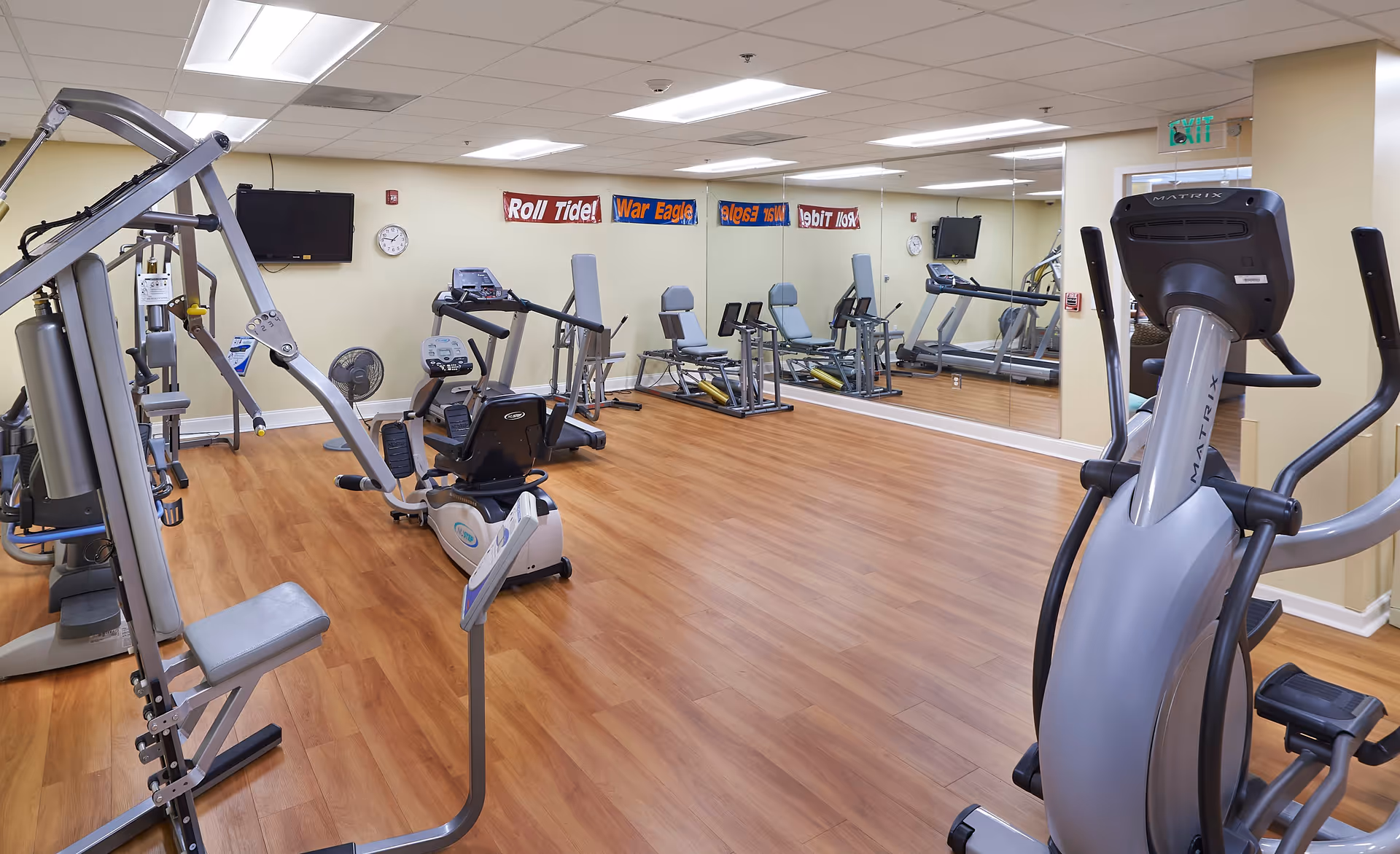 A fitness room with various exercise equipment including a multi-station weight machine, recumbent bike, elliptical machine, and treadmills. The room has wood flooring, mirrored walls, a wall-mounted TV, and banners on the wall that say 'Roll Tide!' and 'War Eagle'.