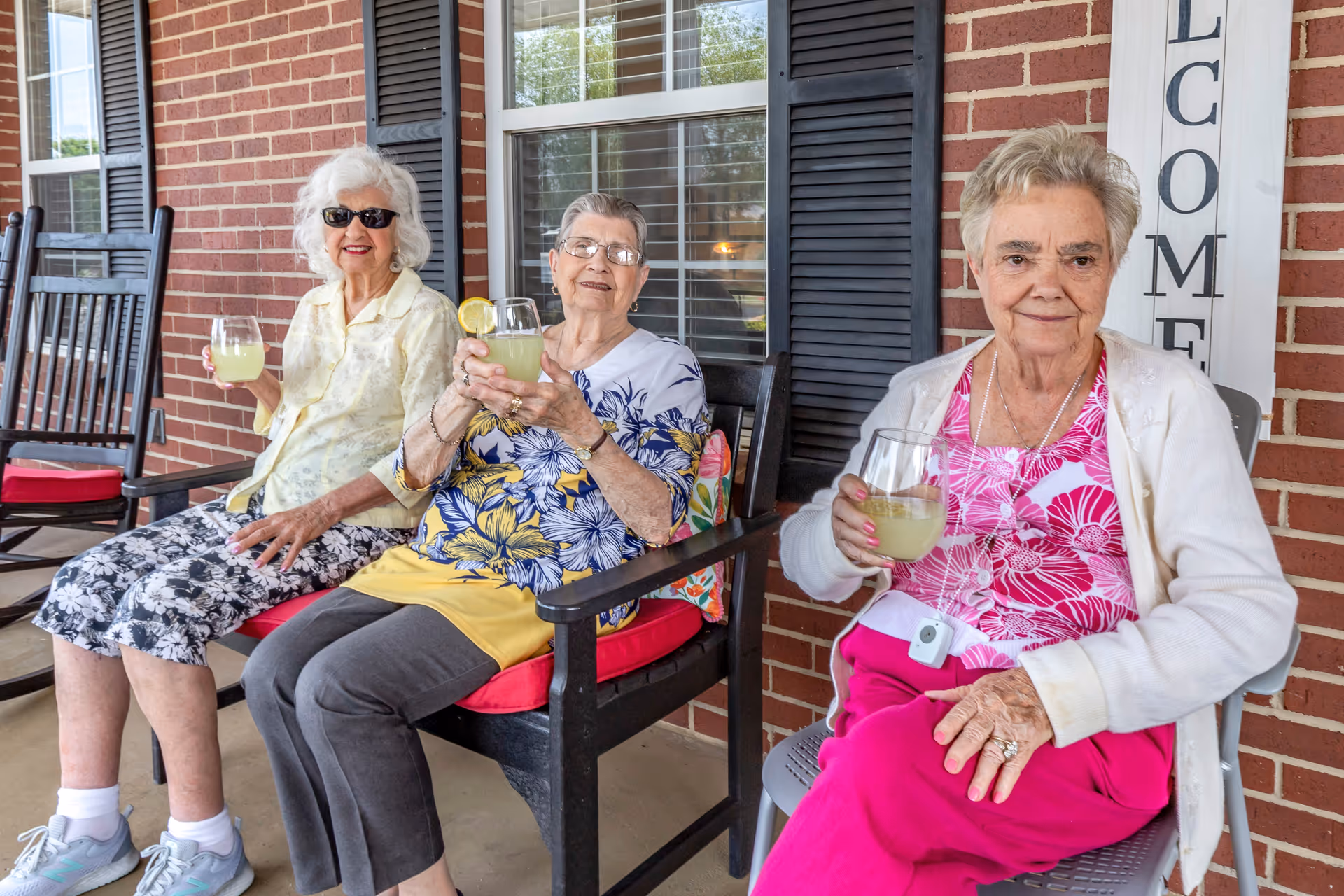 Three elderly women sitting on chairs on a porch with brick walls and windows behind them. They are smiling and holding glasses of lemonade, enjoying a relaxed moment together. One woman is wearing sunglasses, and there is a vertical 'WELCOME' sign on the wall.