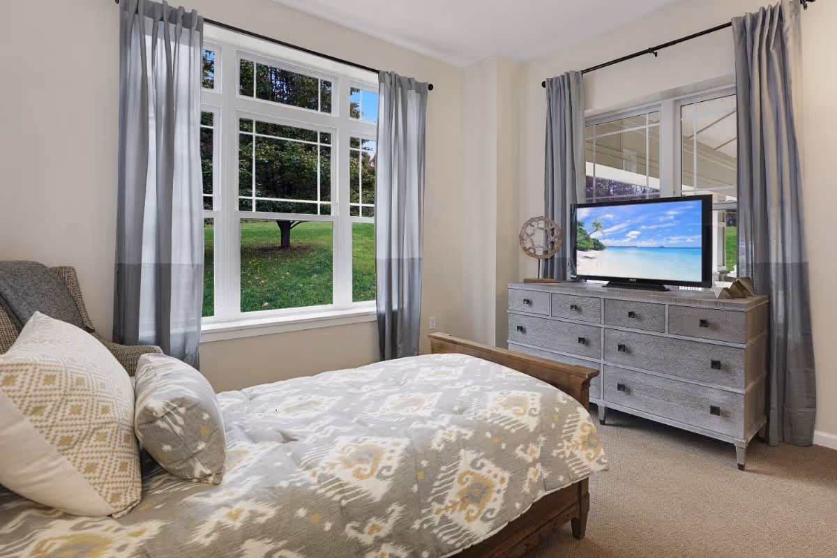 A cozy bedroom with a wooden bed covered in a patterned gray and white comforter and pillows. There are large windows with gray curtains showing a green outdoor view. A gray dresser with multiple drawers holds a flat-screen TV displaying a beach scene, and a decorative object is placed beside the TV.