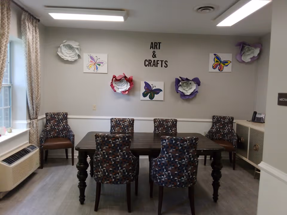 A room with a dark wooden table surrounded by six patterned chairs. The wall behind the table has decorative large paper flowers and butterfly paintings, with the words 'ART & CRAFTS' displayed in black letters. There is a window with patterned curtains on the left and a cabinet with a phone on the right.