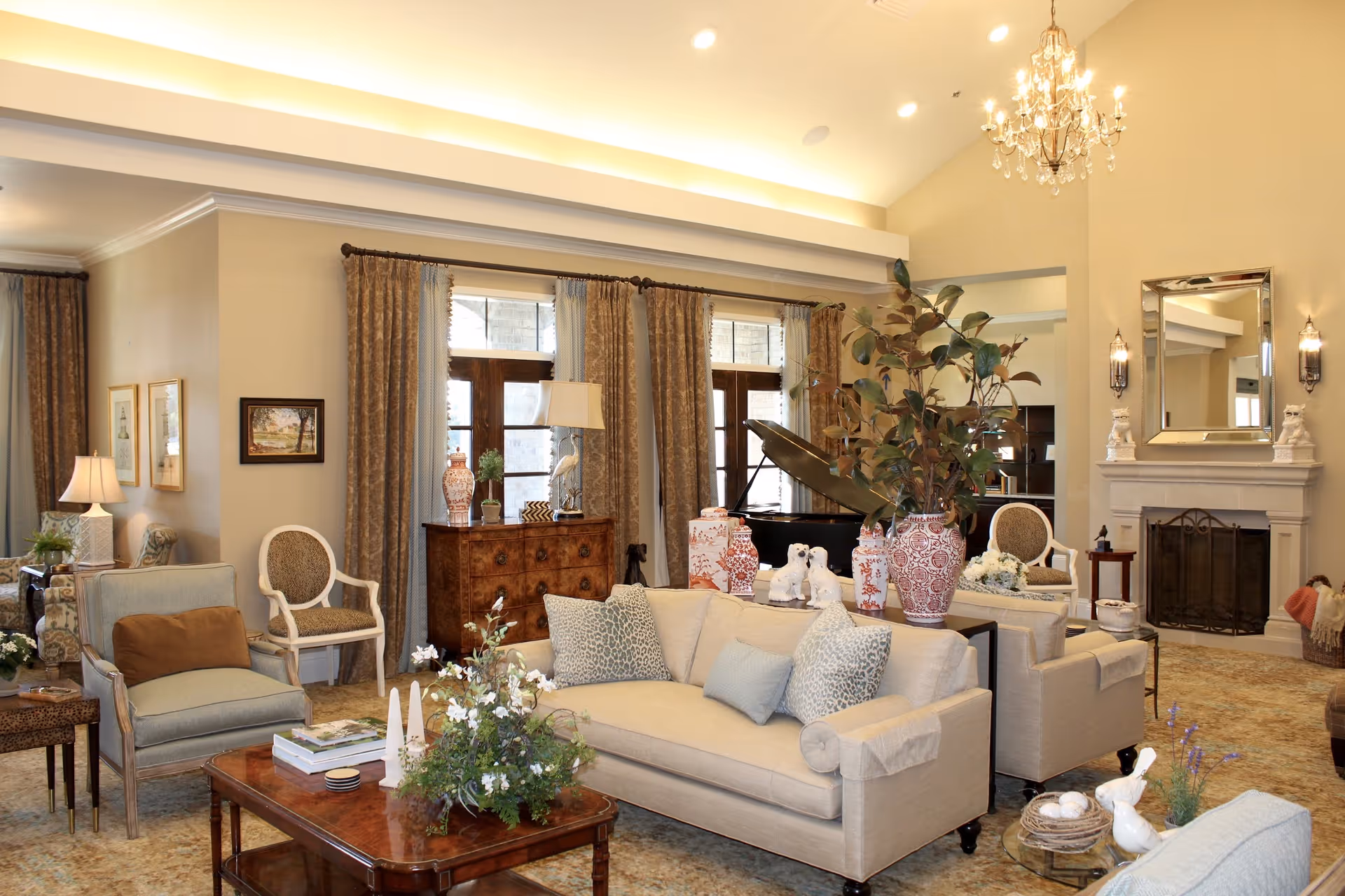 A spacious and elegantly decorated living room in a senior living facility featuring beige sofas with patterned cushions, armchairs, a wooden coffee table with floral arrangements, a grand piano near large windows with heavy curtains, a fireplace with a mirror above it, and a chandelier hanging from the ceiling.
