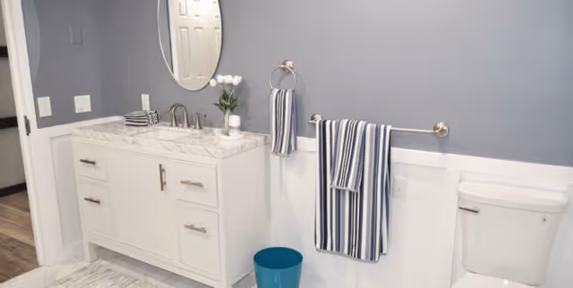 A clean and modern bathroom featuring a white vanity with a marble countertop, a round mirror above the sink, striped blue and white towels hanging on towel racks, a small vase with white flowers, a blue waste bin, and a white toilet against a light blue and white wall.