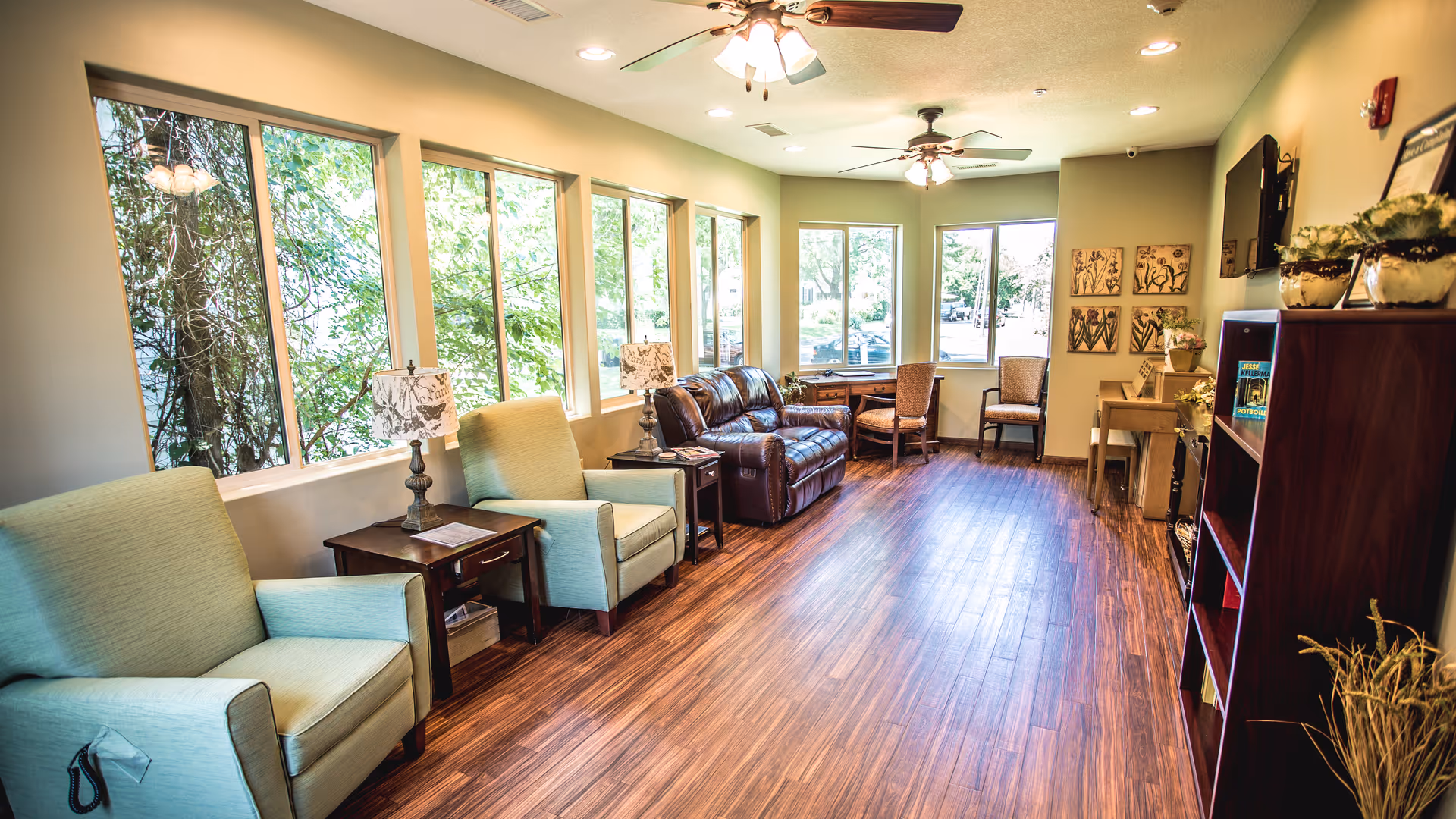 A bright and spacious living room with large windows letting in natural light. The room features two light green armchairs with a small wooden side table and lamp between them, a brown leather sofa, two wooden chairs near a window, a wooden bookshelf with decorative items, and a wooden floor. Ceiling fans with lights are mounted on the ceiling.