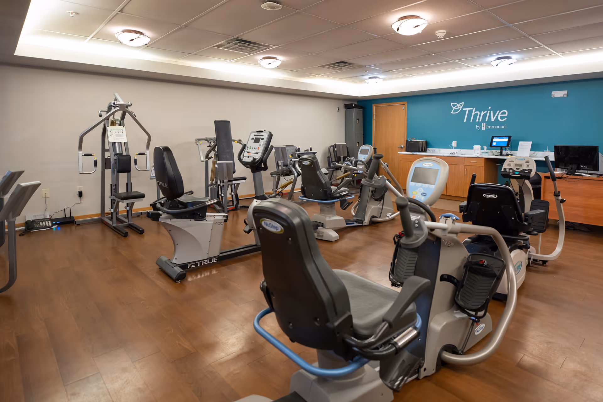Interior view of a fitness room with various exercise machines including recumbent bikes and strength training equipment. The room has wooden flooring, a teal accent wall with the text 'Thrive by Immanuel', and a counter with a computer and other items.
