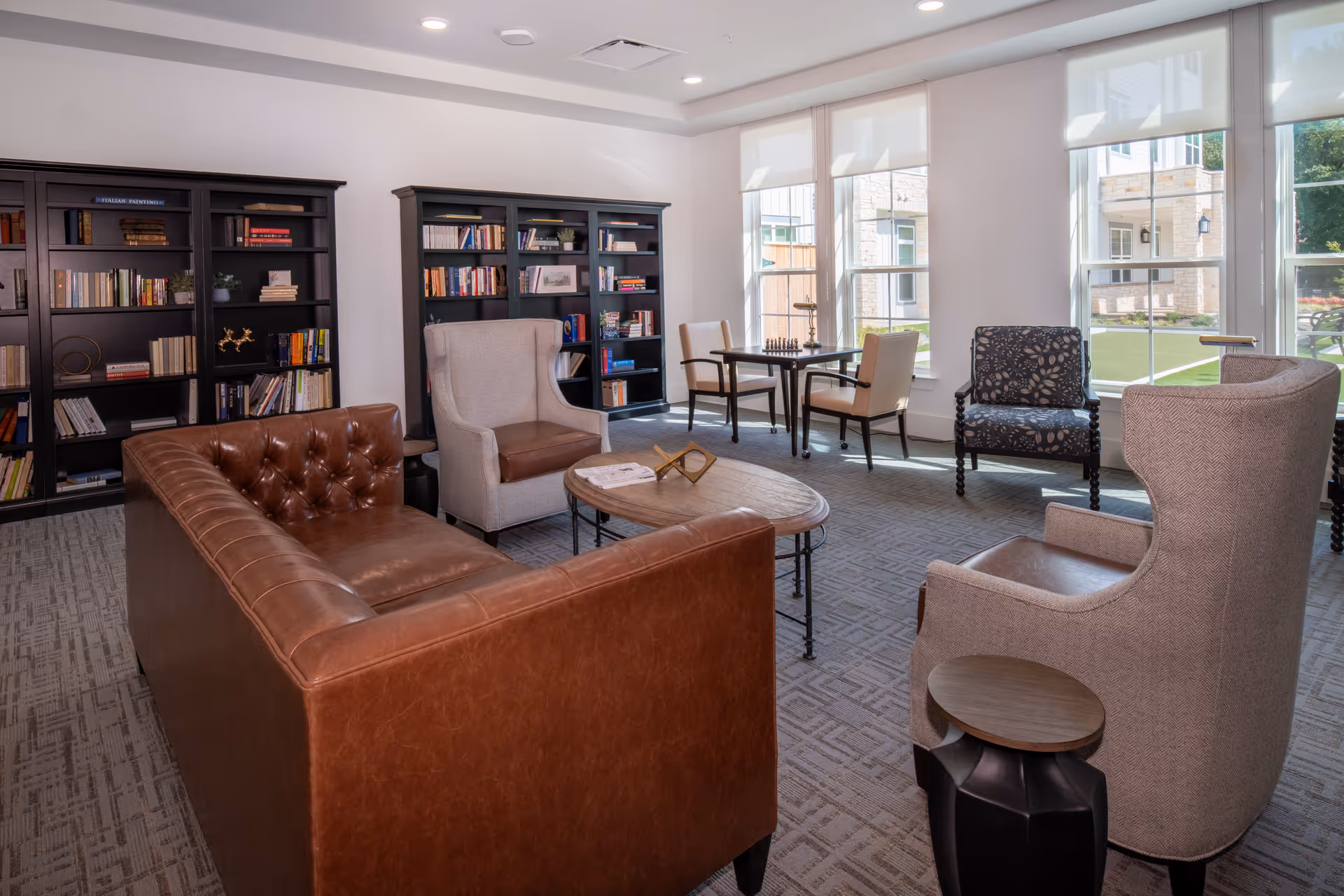 A bright and cozy common area with large windows letting in natural light. The room features a brown leather sofa, two beige armchairs with leather cushions, a patterned armchair, and a wooden coffee table. In the background, there are two black bookshelves filled with books and decorative items. Near the windows, there is a small table with two chairs and a chessboard on top. The room has a carpeted floor and white walls.