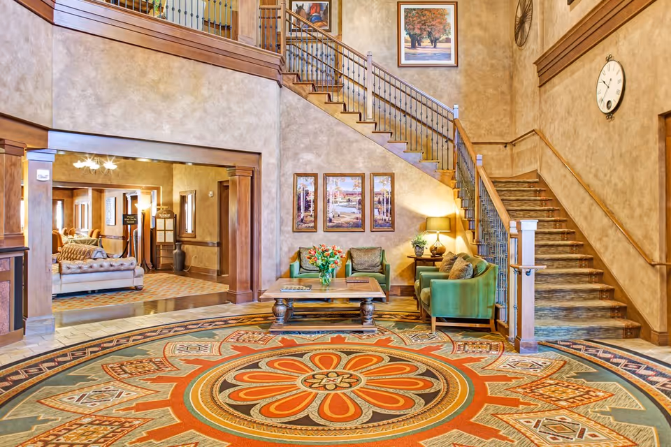 A spacious and warmly decorated senior living facility common area with a large circular patterned rug in the center, green upholstered chairs around a wooden coffee table with a vase of flowers, a staircase with wooden handrails and iron balusters leading to an upper floor, and framed artwork on the walls. In the background, there is another seating area with a sofa and additional chairs.