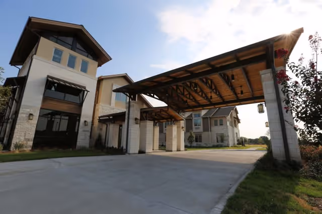 Exterior view of The Healthcare Resort of Plano showing a covered driveway entrance with wooden beams and stone pillars, adjacent to a multi-story building with large windows and a sloped roof under a partly cloudy sky.