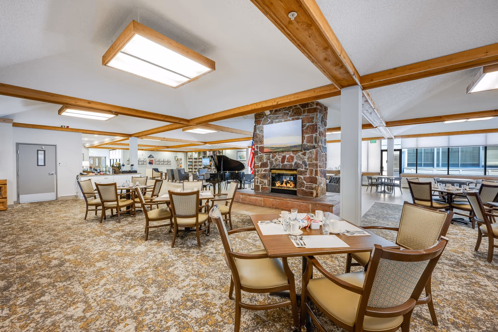 A spacious dining room with multiple tables and chairs arranged for meals. The room features a stone fireplace with a painting above it, wooden ceiling beams, and large windows letting in natural light. There is a grand piano near the fireplace and a carpeted floor with a patterned design.