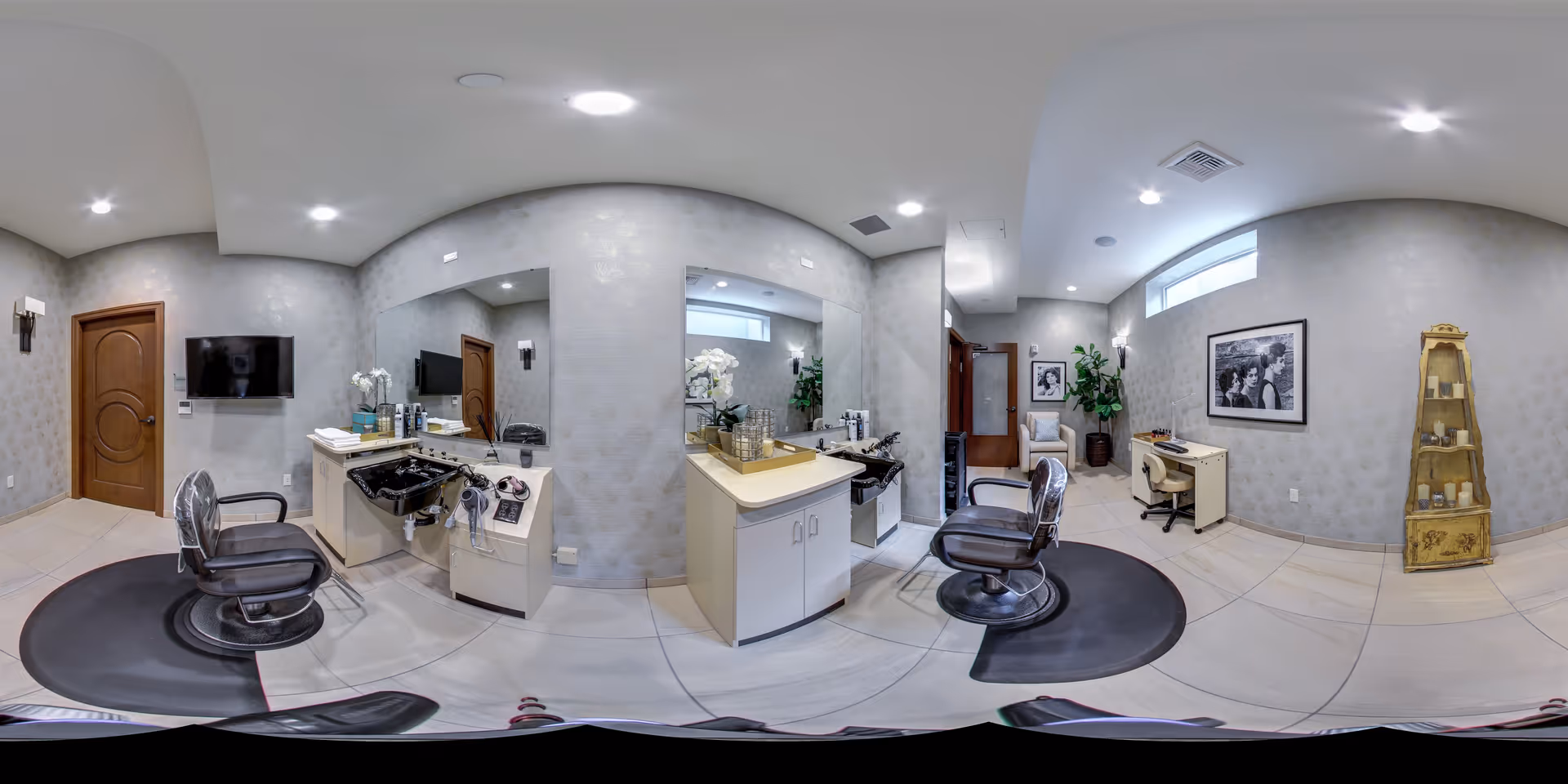 A well-lit salon room with two black salon chairs positioned in front of sinks and mirrors. The room has light-colored tiled floors and gray patterned walls. There is a wooden door, a wall-mounted TV, a small desk with a chair, framed black and white photos on the wall, a tall decorative shelving unit, and some potted plants. The ceiling has recessed lighting.
