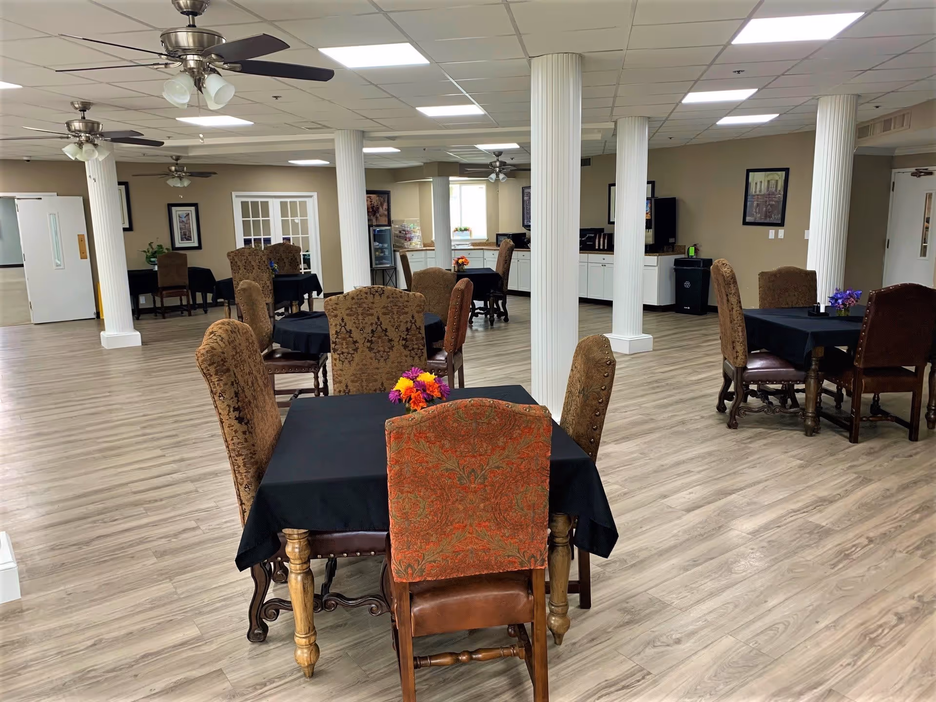 A spacious dining room in a retirement community with several tables covered in black tablecloths and surrounded by upholstered chairs. The room features light wood flooring, white columns, ceiling fans with lights, and framed artwork on beige walls. There is a counter area with cabinets and a coffee machine in the background.