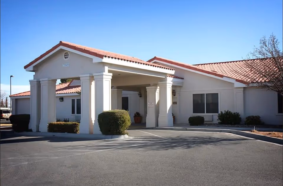 Exterior view of a single-story senior living facility building with a covered entrance supported by columns, red-tiled roof, and surrounding bushes under a clear blue sky.