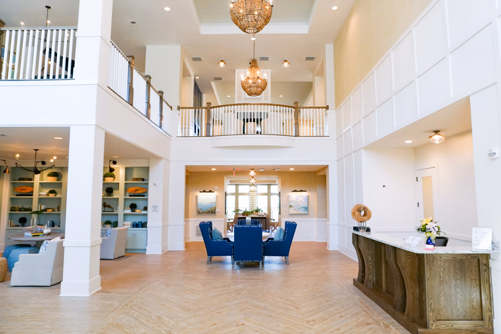 Spacious and bright senior living facility common area with high ceilings, two large chandeliers, a wooden reception desk with flowers, blue cushioned chairs around a table, and a second-floor balcony overlooking the space.