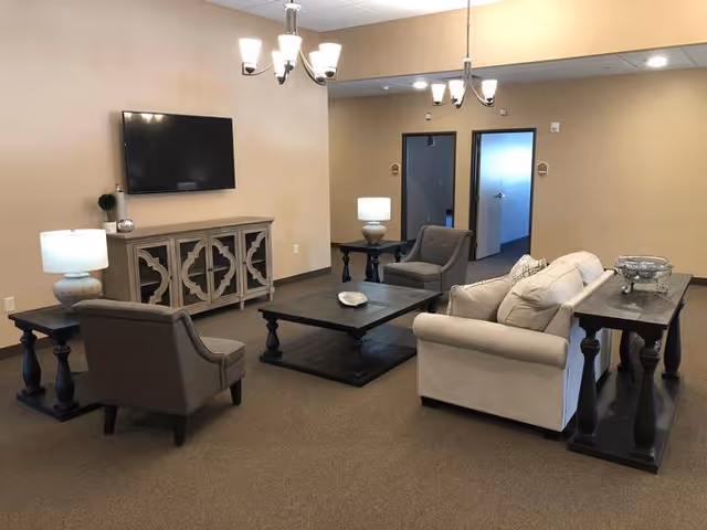 A cozy living room area with beige walls and carpeted floor. The room features a beige sofa with cushions, two gray armchairs, a dark wooden coffee table, and matching side tables with lamps. A decorative cabinet with a mounted flat-screen TV is against one wall. Two chandeliers hang from the ceiling, and there are two doorways visible in the background.