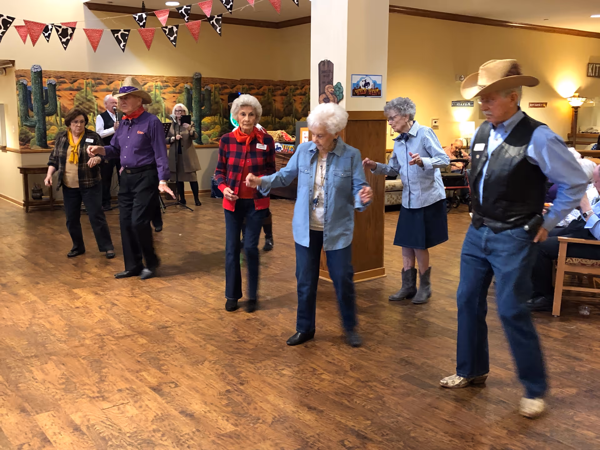 A group of elderly people dressed in western attire are line dancing in a room with wooden floors and western-themed decorations. In the background, a small band is playing music. The room has a warm, inviting atmosphere with yellow walls and festive bunting hanging from the ceiling.