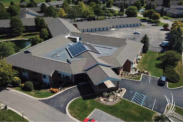 Aerial view of a single-story assisted living facility with a large roof featuring solar panels. The building is surrounded by parking lots, landscaped greenery, and nearby residential houses. The facility has a covered entrance and multiple garage doors at the back.