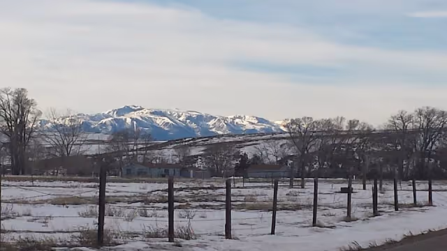 Snowy rural field with a wooden fence, a few houses and leafless trees, and snow-capped mountains under a pale sky.