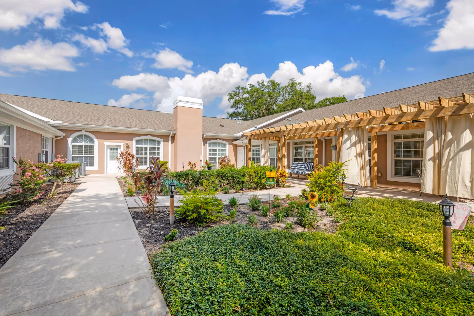 Sunlit courtyard with a paved walkway, landscaped gardens, and a single-story building featuring a wooden pergola and arched windows.