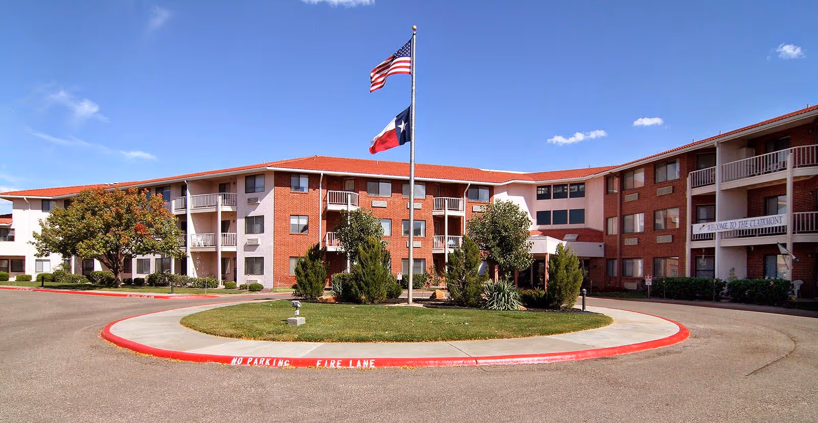 Exterior view of a three-story senior living facility with red brick and white walls, balconies, and a circular driveway with a grassy island in the center featuring two flagpoles flying the American and Texas flags under a clear blue sky.