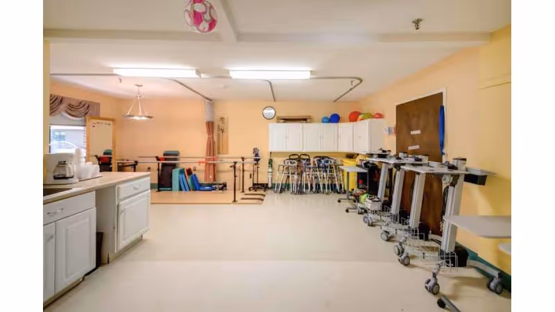A rehabilitation therapy room with parallel bars for walking exercises, several walkers lined up against the wall, exercise balls on top of white cabinets, and various therapy equipment. The room has beige walls and a white ceiling with fluorescent lighting.