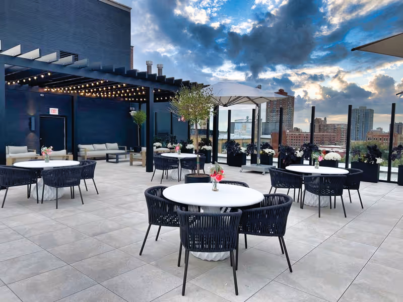 Rooftop patio with round white tables, black woven chairs, a pergola with string lights, planters and city skyline under a cloudy sky.