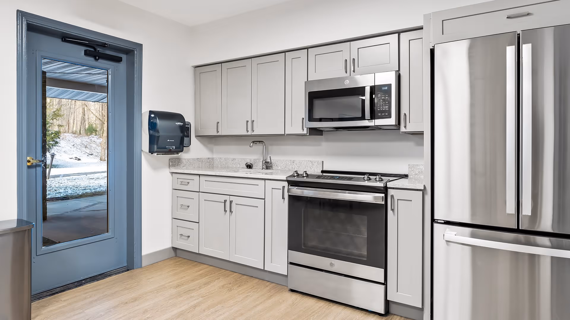 Modern kitchen area with light gray cabinets, a stainless steel oven with a microwave above it, a stainless steel refrigerator, a sink, and a blue door leading outside with a view of a snowy wooded area.