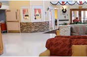 Interior view of a nursing and rehabilitation center common area with a red patterned sofa in the foreground, a brick half-wall, windows with red curtains, and holiday decorations including garlands and a wreath.