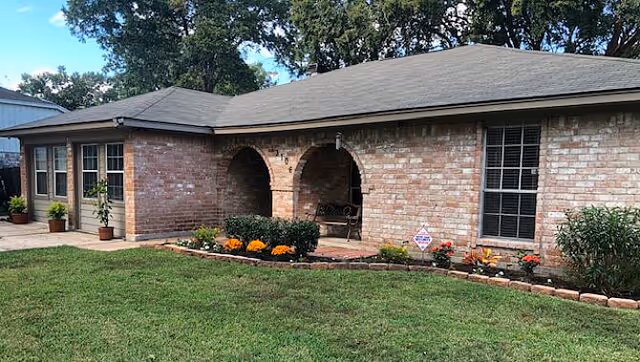 Exterior view of a single-story brick building with a gray shingled roof, featuring two arched entryways and several windows. The front yard has a well-maintained green lawn and a flower bed with orange and red flowers along the building's foundation.