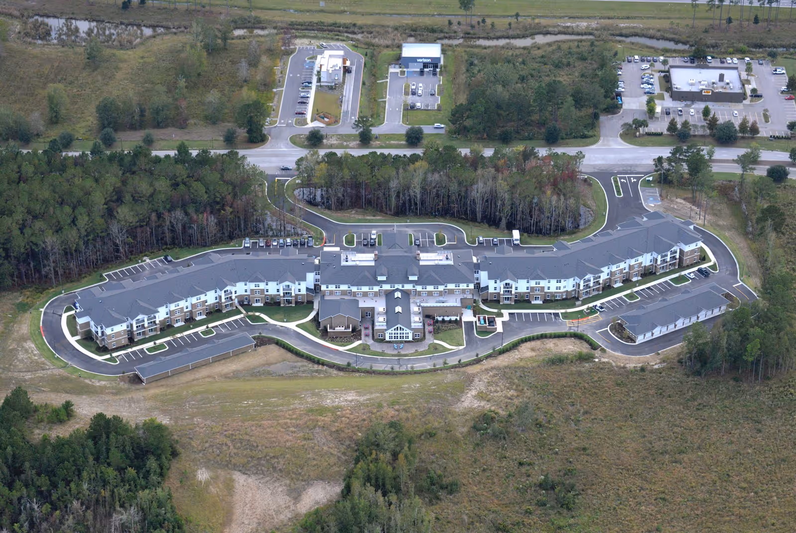 Aerial view of a long, curved multi-story retirement community building with parking, driveways, surrounding trees and open land.
