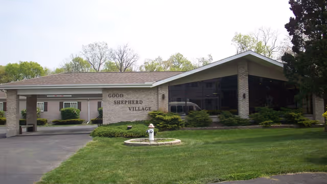 Exterior front view of Good Shepherd Village building with a covered entrance, large windows, and surrounding greenery including bushes and trees.
