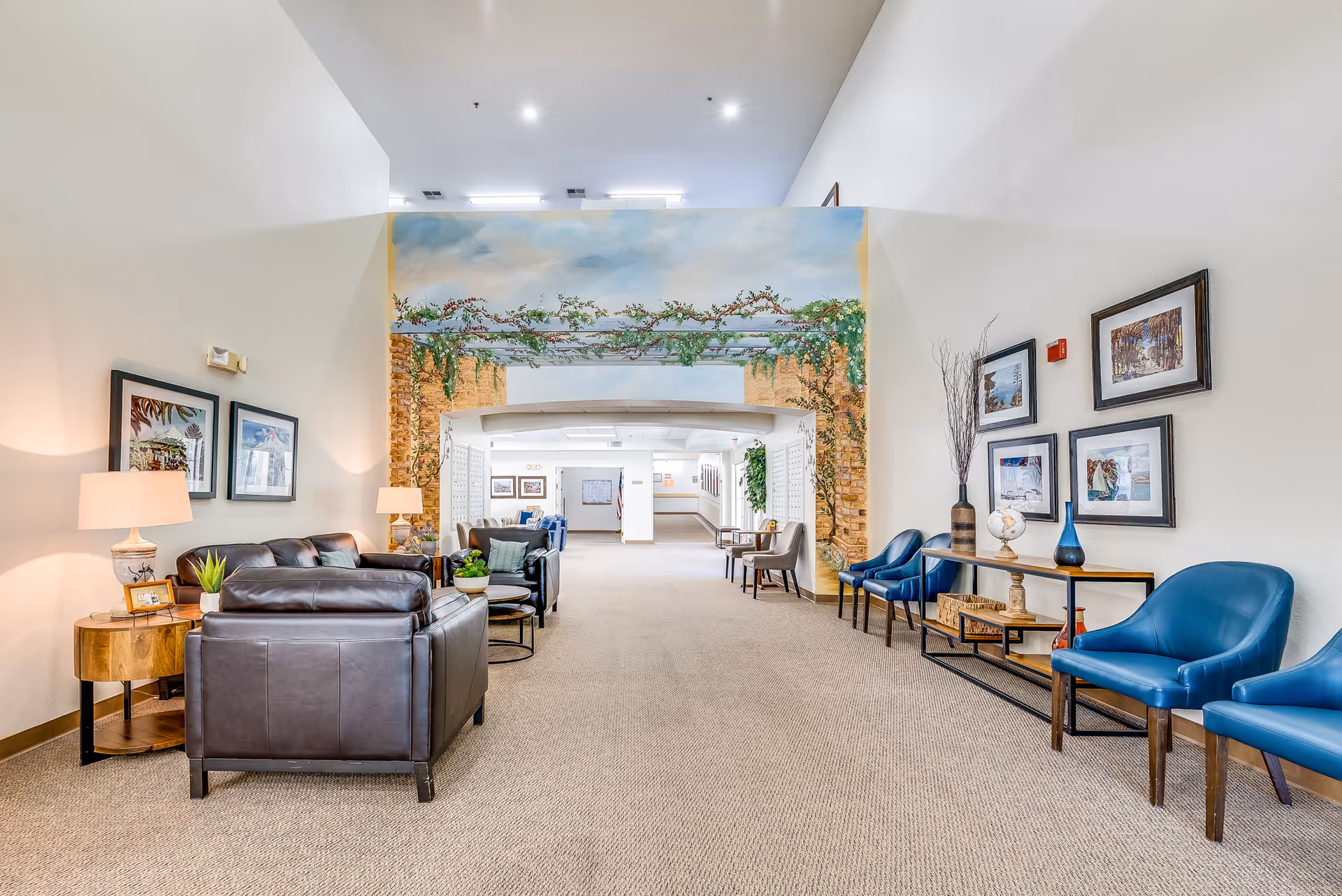 Spacious senior living facility common area and lounge with leather sofas, blue chairs, side tables and a decorative mural arch over a carpeted hallway.
