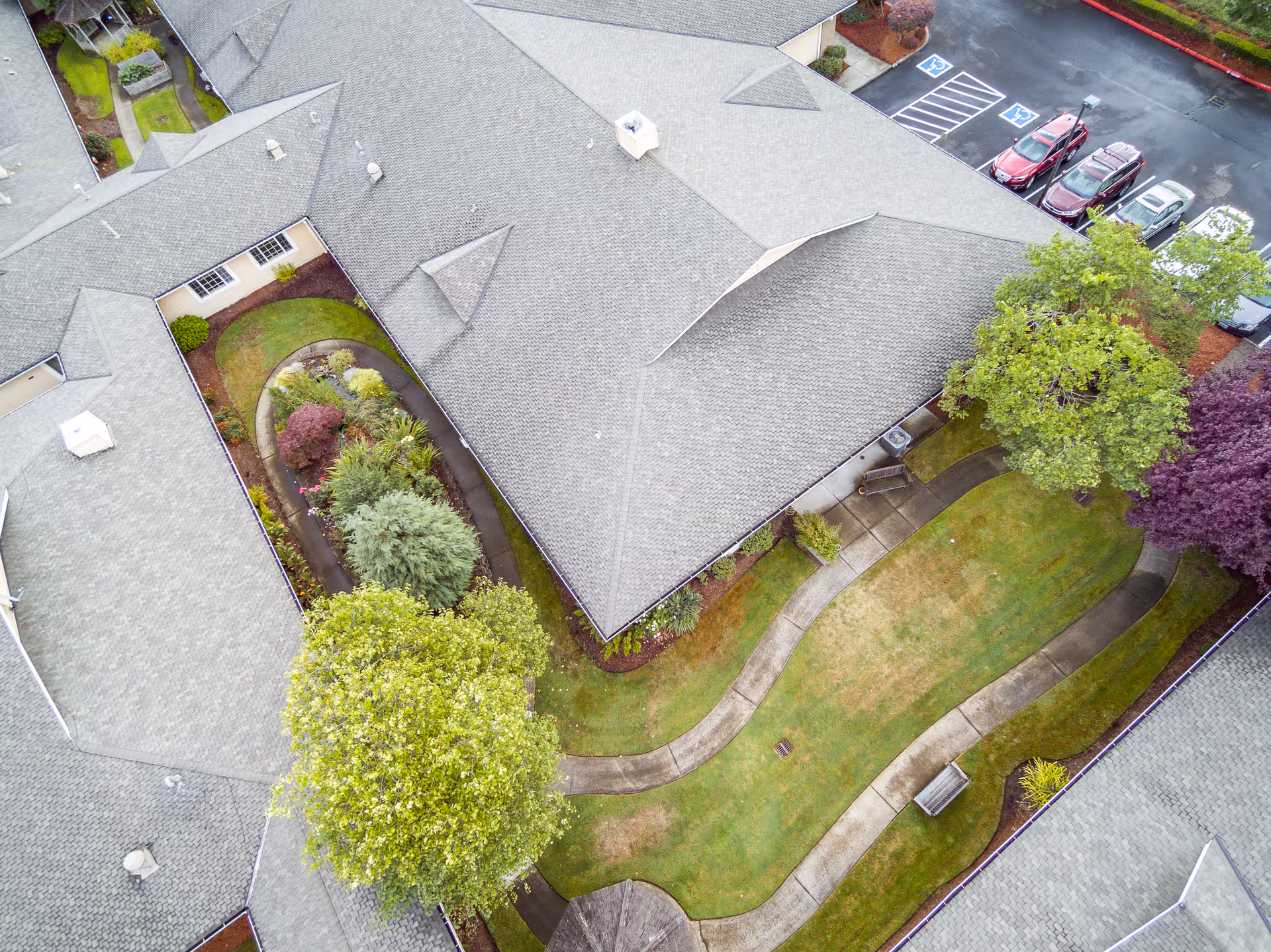 Aerial view of a senior living facility showing a courtyard with winding paved walkways, green lawns, trees, and landscaped garden beds surrounded by building rooftops and a parking area with several cars.