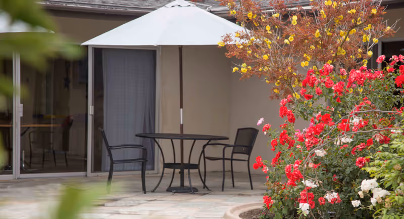 Outdoor patio area with a round black metal table and two matching chairs under a large white umbrella. The patio is adjacent to a building with sliding glass doors. There are vibrant red, white, and yellow flowers and green foliage in the foreground.