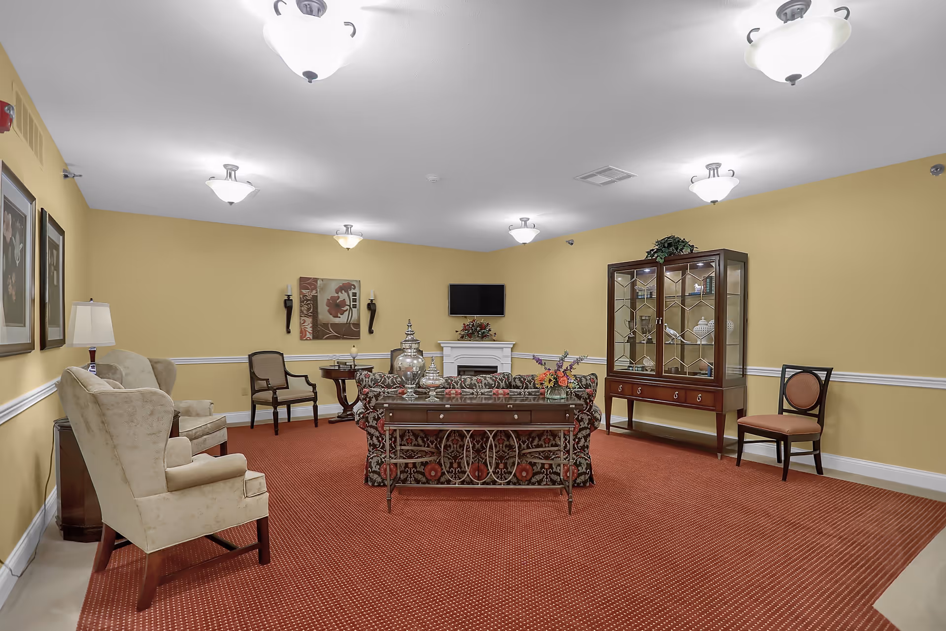 A cozy living room with yellow walls and red carpet. The room features a floral patterned sofa, two beige armchairs, a wooden display cabinet with glass doors, a small table with chairs, a wall-mounted TV above a white fireplace, and decorative wall art and lighting fixtures.