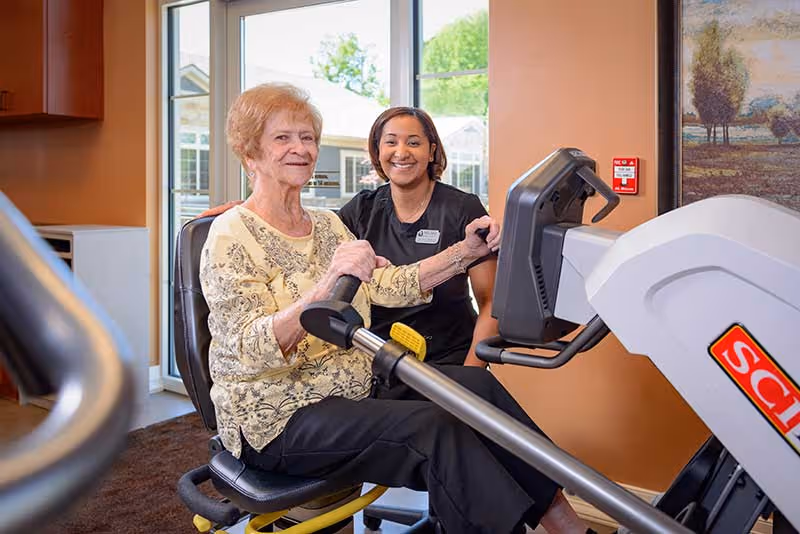 An elderly woman using a seated exercise machine in a fitness room, assisted by a smiling staff member wearing a black uniform. The room has large windows with a view of greenery outside and warm-colored walls with a framed painting.