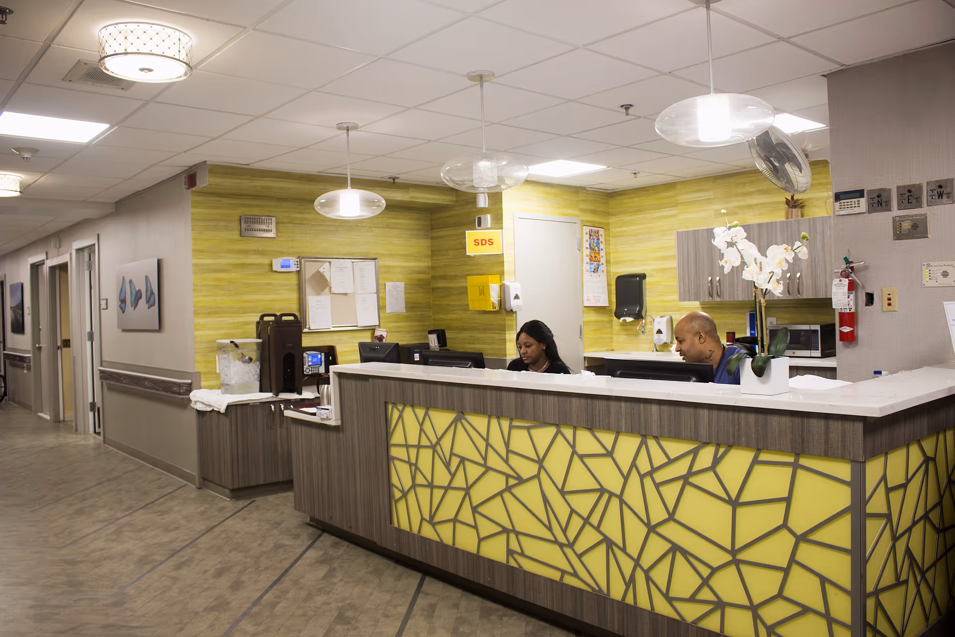 Reception desk area inside a nursing facility with two staff working behind a patterned counter and a hallway to the left.