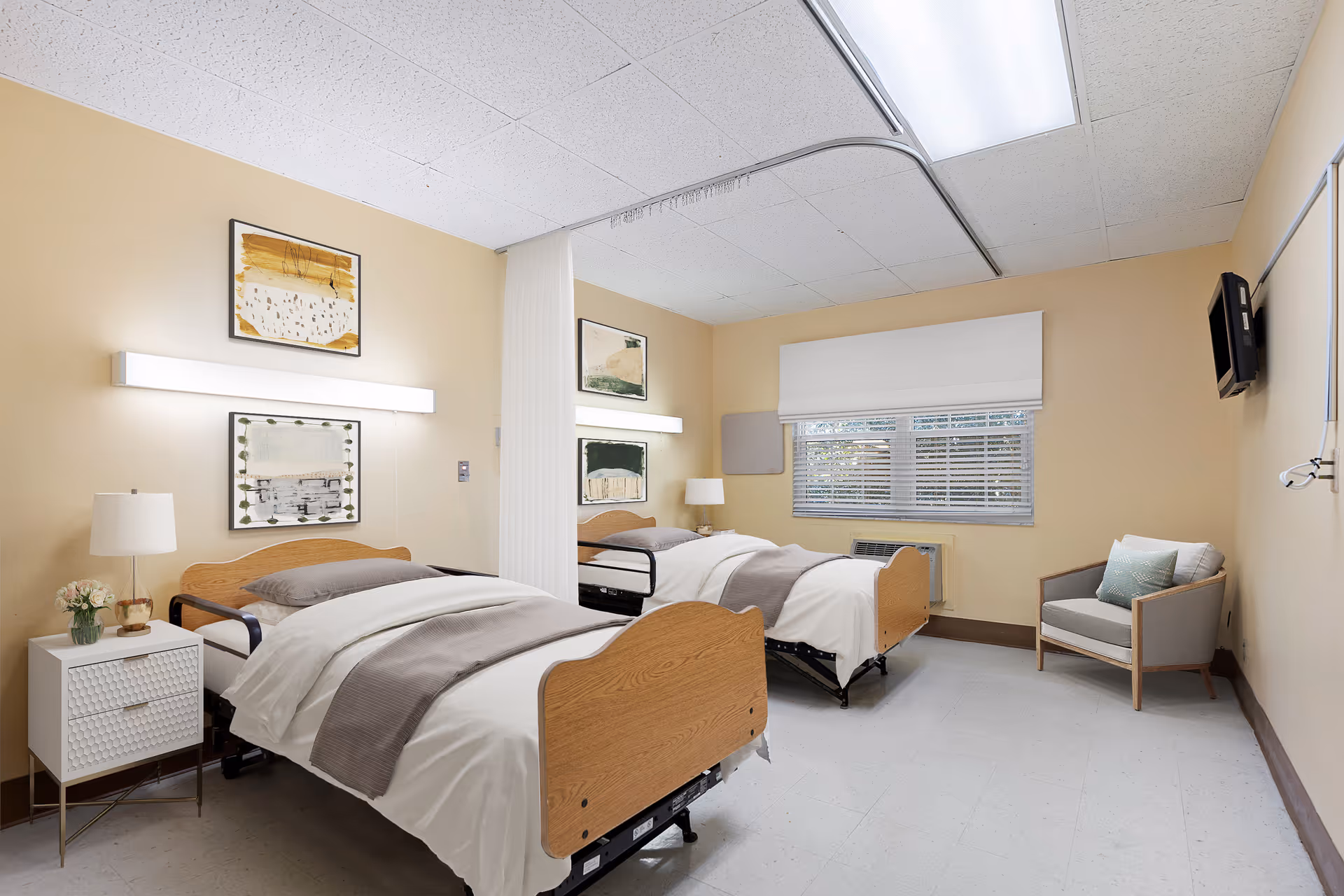 A shared patient room with two adjustable hospital beds, bedside tables, artwork on the walls, a chair, and a window with blinds.