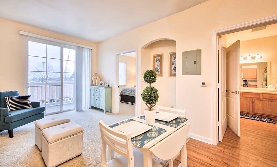 Bright and cozy living area in a senior living facility with a small dining table set for two, a blue armchair with a striped pillow, two beige ottomans, a light green cabinet, and sliding glass doors leading to a balcony. The room has beige walls and carpeted floors. A bedroom is visible through an open doorway, and a bathroom with wooden cabinets and a large mirror is seen through another open door.