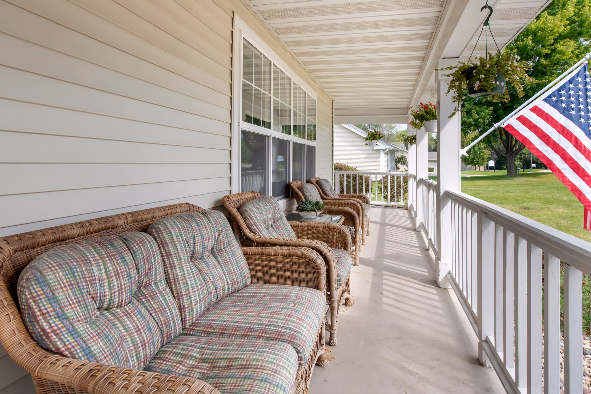 Covered front porch with wicker seating, hanging plants, and an American flag overlooking a grassy lawn.