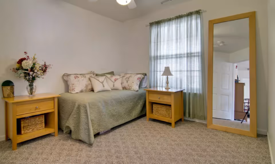 A cozy bedroom with a single bed covered in a green quilt and floral pillows. There are two wooden nightstands on either side of the bed, one with a vase of flowers and the other with a small lamp. A large wooden framed mirror stands against the wall near a window with sheer curtains, and the floor is carpeted.