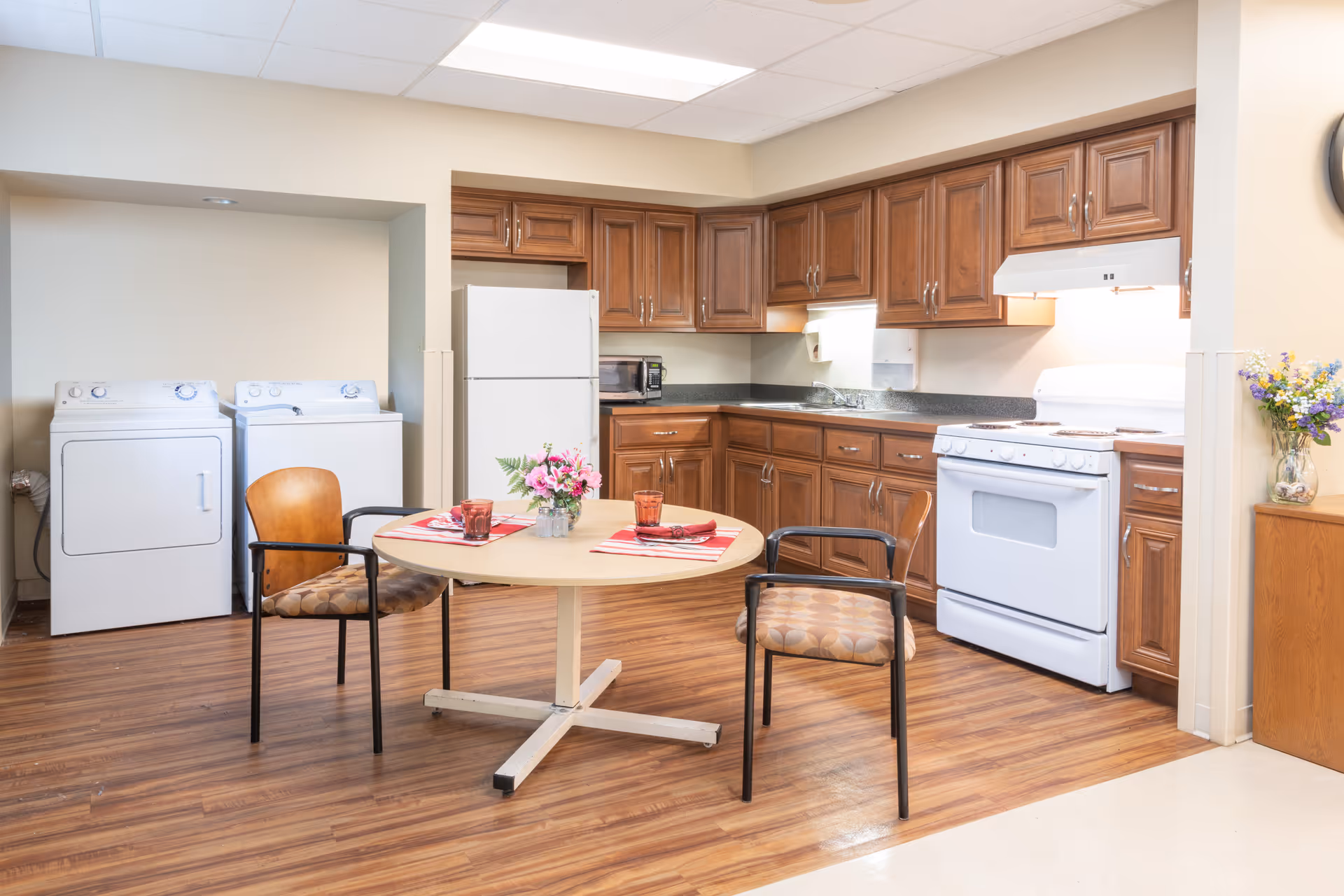 A communal kitchen and dining area with wooden cabinets, a round table with chairs, refrigerator and stove, and a washer and dryer.
