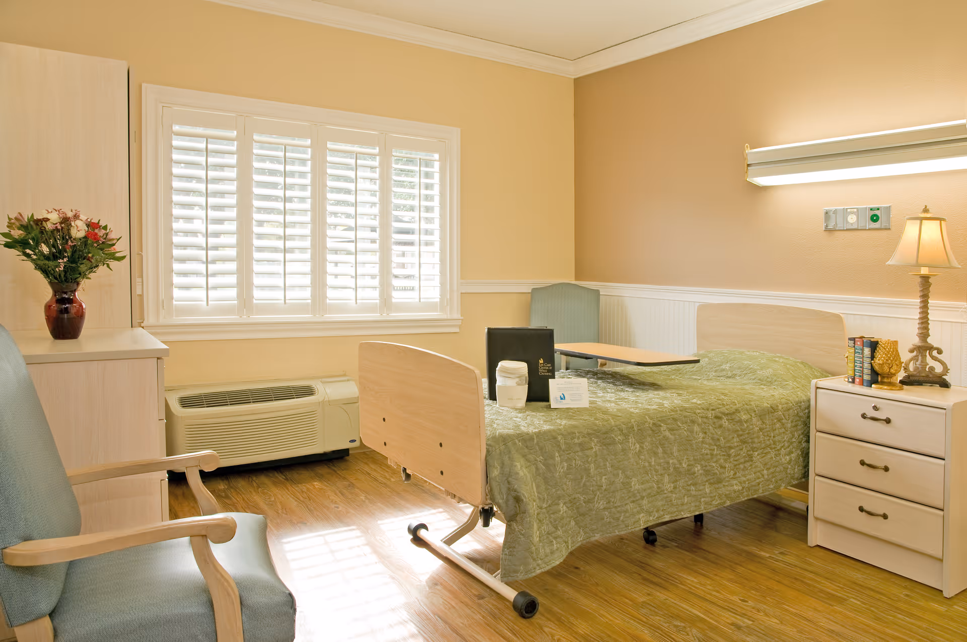 A well-lit senior living bedroom with a hospital-style bed covered in a green quilt, a bedside table with a lamp and books, a comfortable armchair, a small table on wheels, a cabinet with a vase of flowers, and a large window with white shutters letting in natural light.