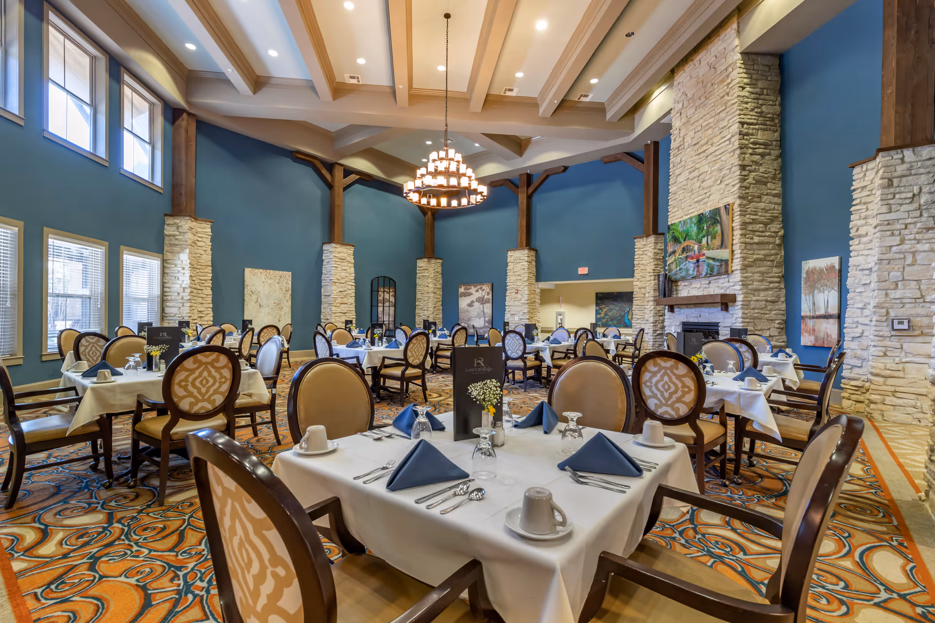 A spacious dining room with high ceilings and large windows. The room features multiple tables covered with white tablecloths, each set with blue folded napkins, cups, glasses, and silverware. The walls are painted blue with stone pillars and decorative paintings. A large chandelier hangs from the ceiling, and the floor is covered with a patterned carpet.