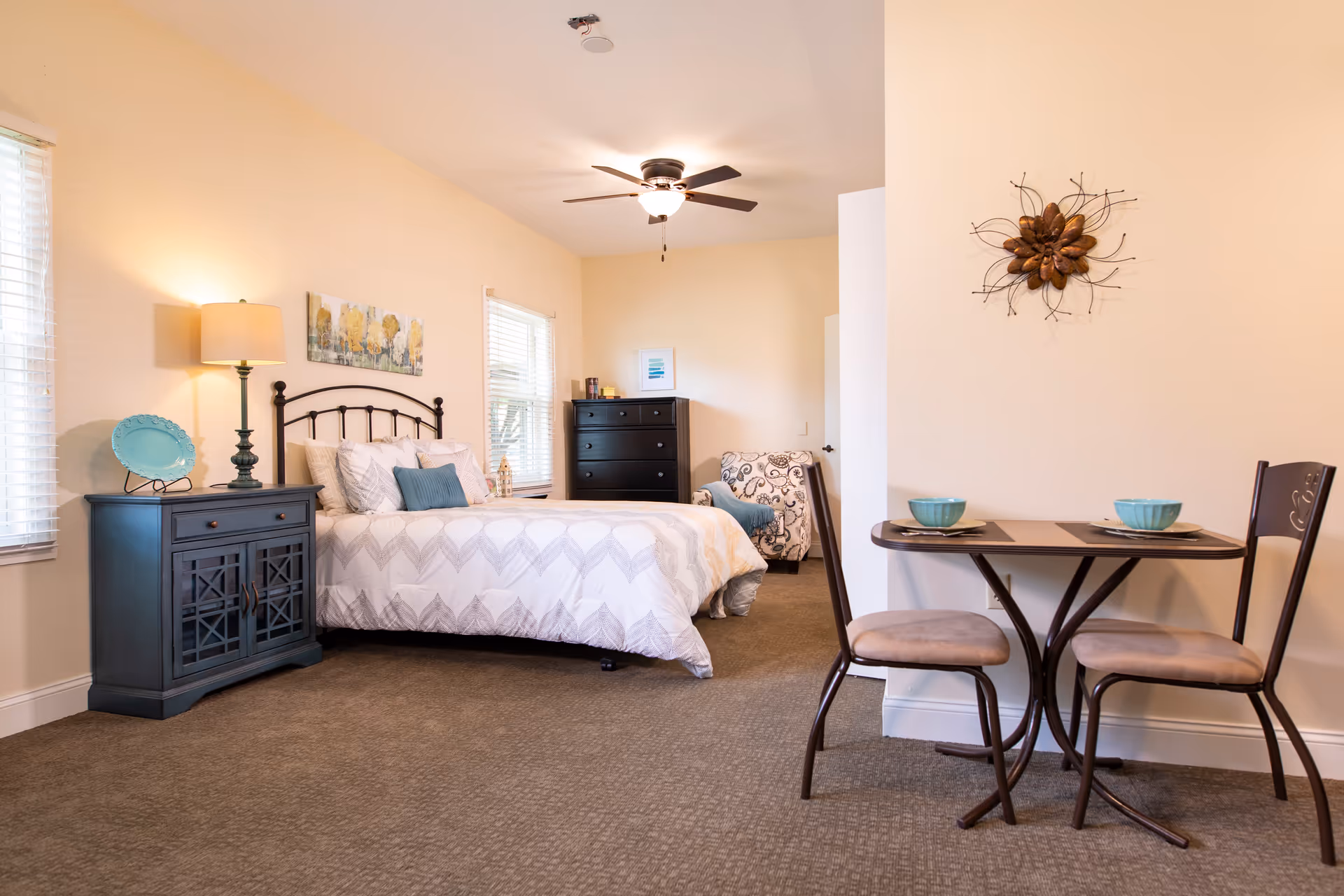 Well-lit studio bedroom with a bed, nightstand and lamp, dresser, armchair, and a small two-person dining table.